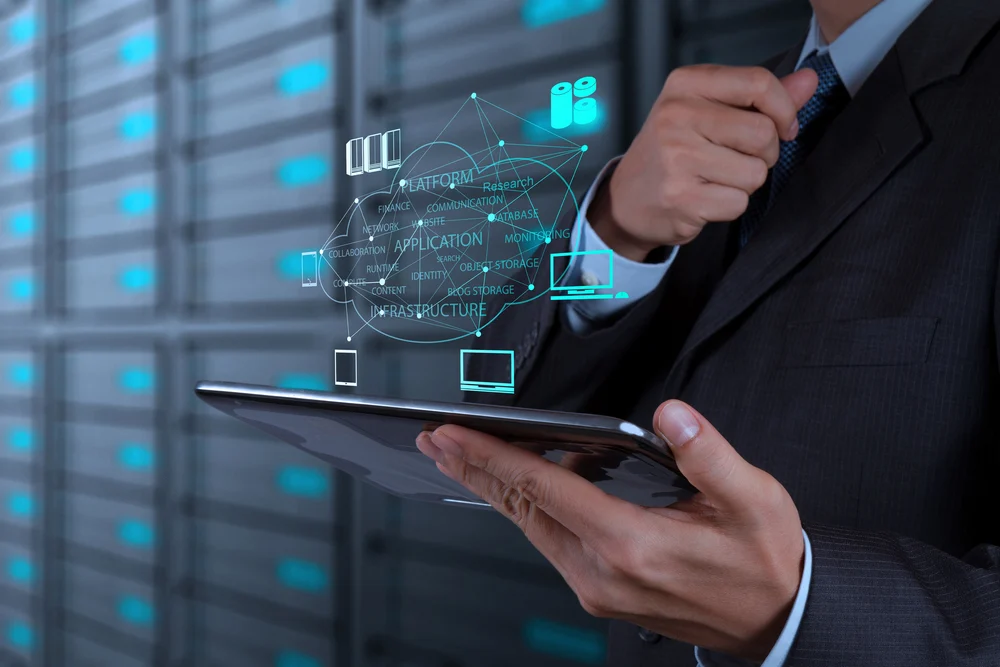 A professional holding a tablet that projects a holographic cloud computing network, with glowing blue server racks in AI data centers in the background.